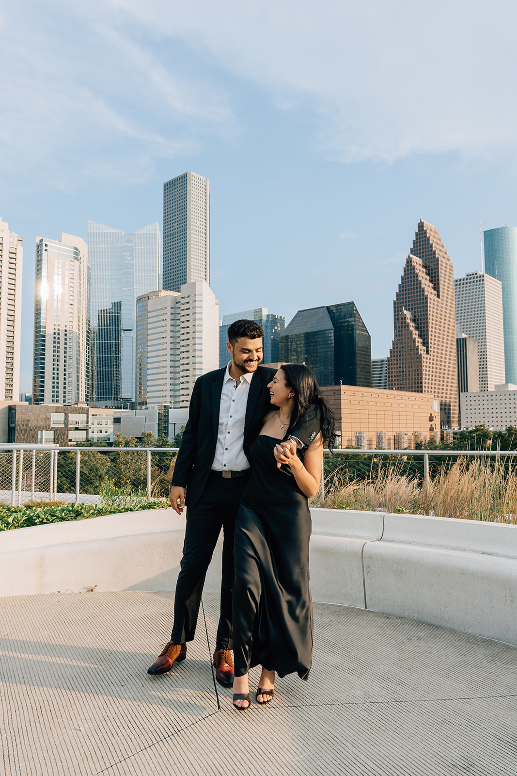 Bride and groom walk hand in hand on the rooftop of POST Houston during golden hour, smiling with the downtown Houston skyline in the background. Captured by a Houston wedding photographer.