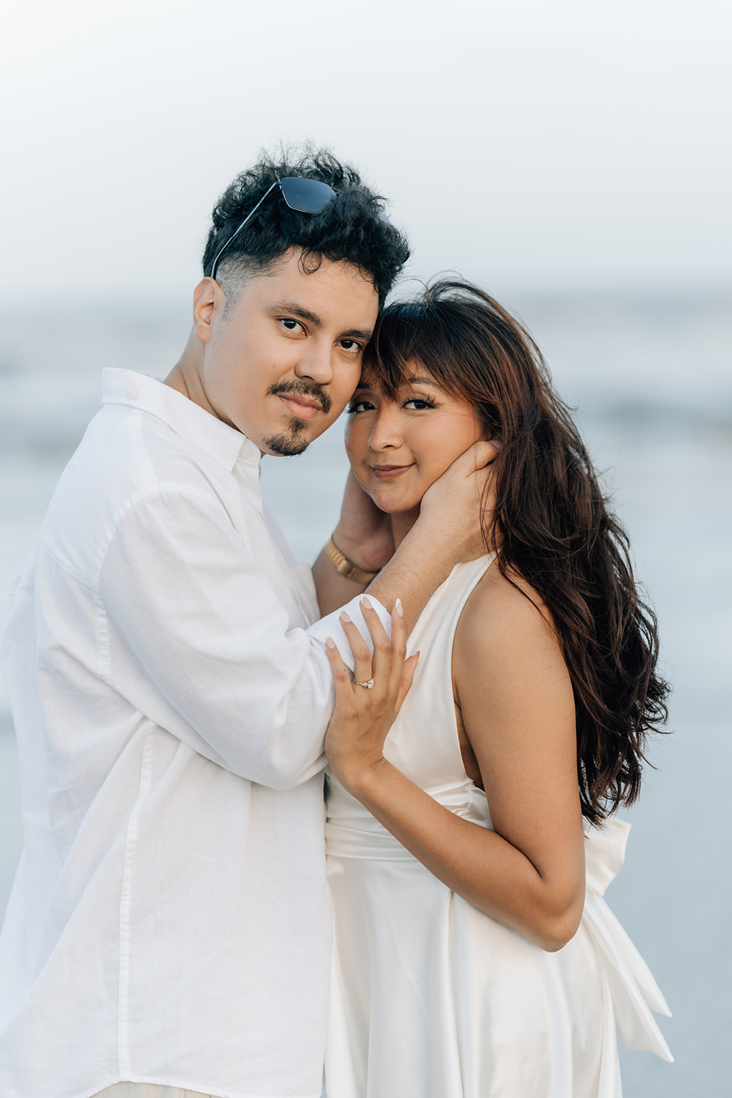 A couple standing close together on the beach during their engagement session, gently holding each other and smiling softly at the camera, photographed in soft coastal light.