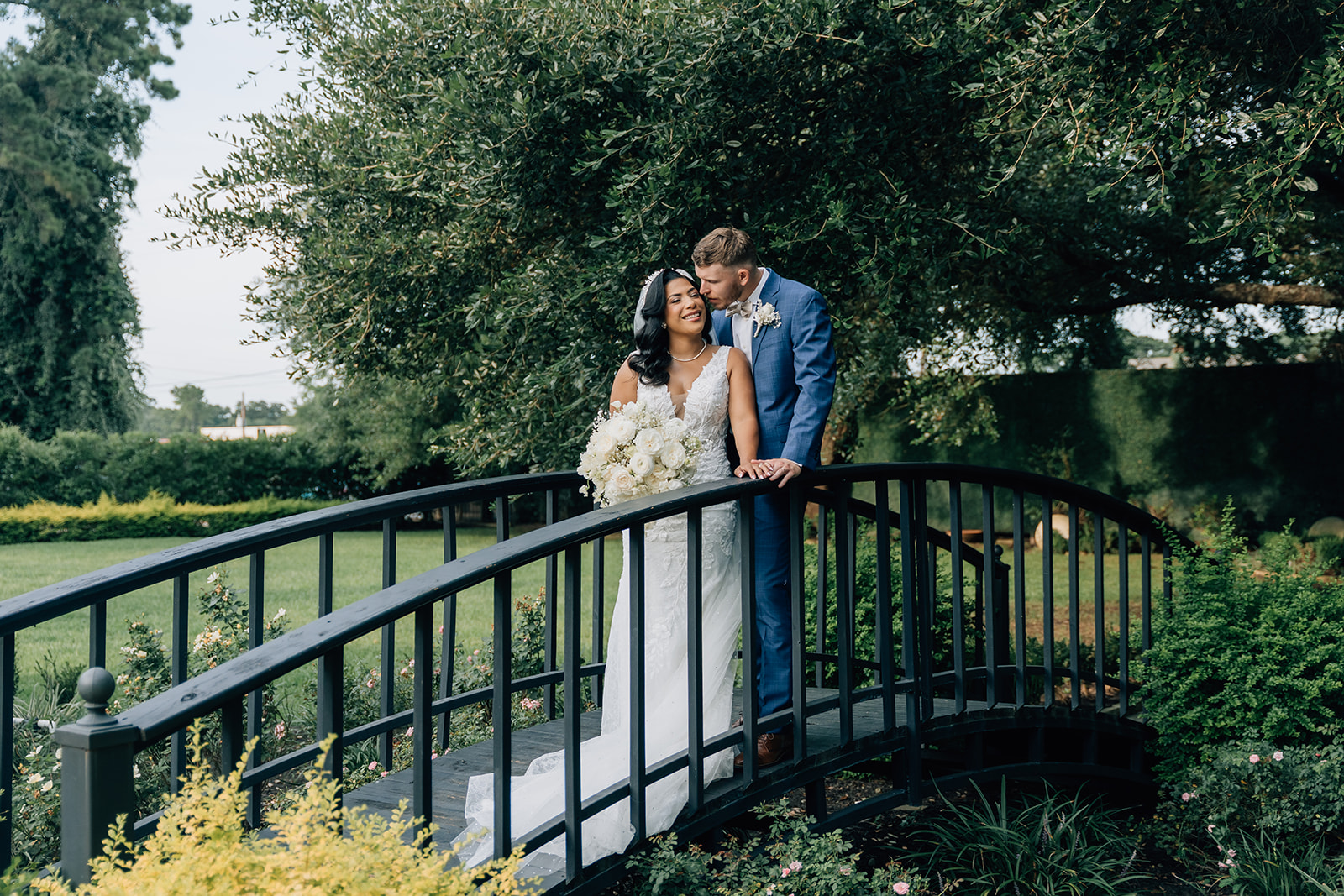 Bride and groom sharing a romantic moment on a black bridge surrounded by greenery at The Pecan Orchard wedding venue near Houston, captured by Katia G Photography.