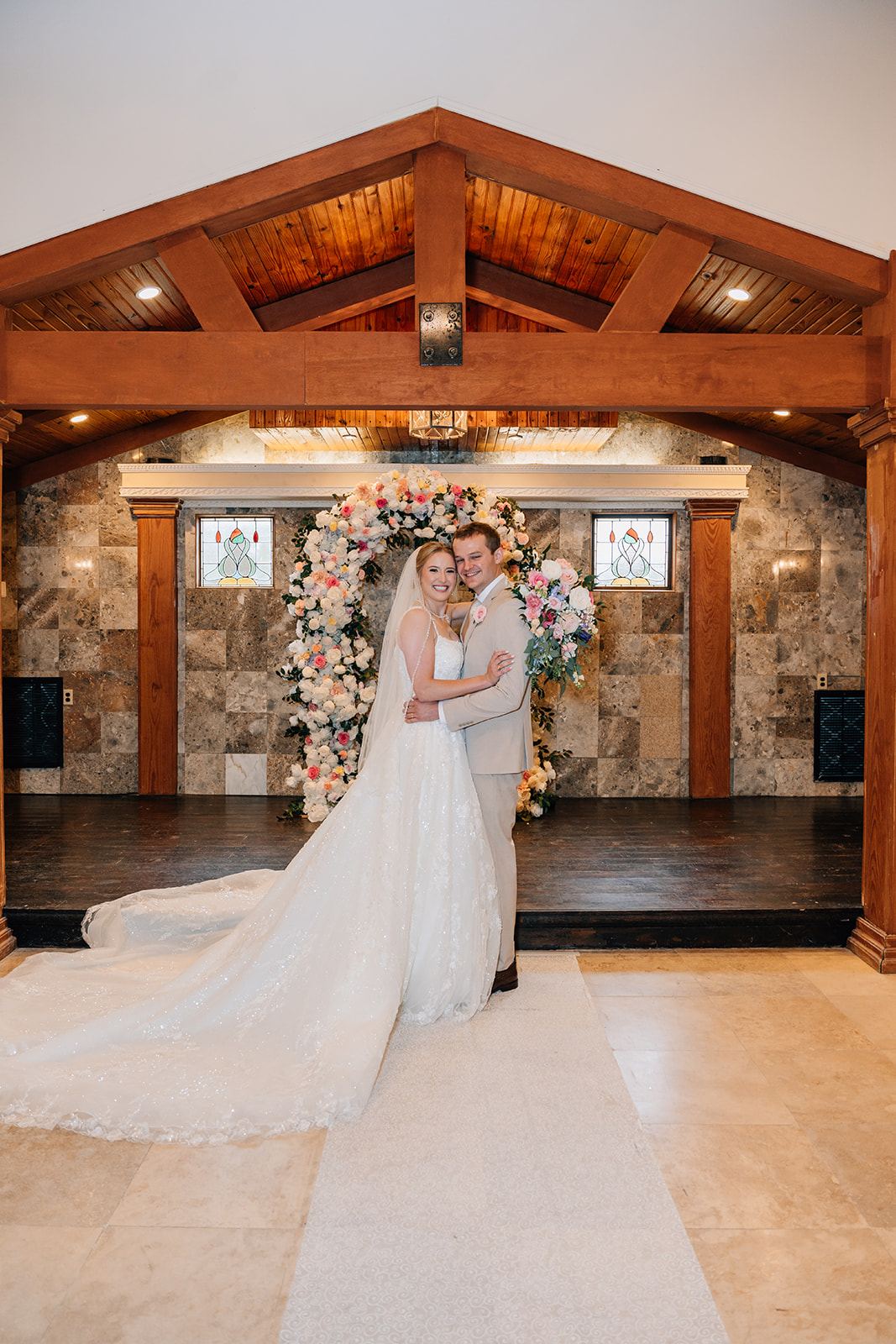A bride and groom stand smiling and embracing at their indoor wedding ceremony at Safari Texas Ranch. The bride wears a long, flowing white gown with a train and veil, while the groom is dressed in a light-colored suit. Behind them is a large circular floral arch filled with pastel flowers, flanked by stained-glass windows and warm wooden beams overhead.