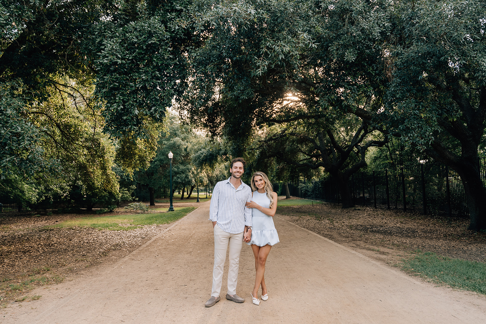 Bride-to-be and groom-to-be standing on a tree-lined path during their Houston engagement session at sunset, captured by a Houston wedding photographer