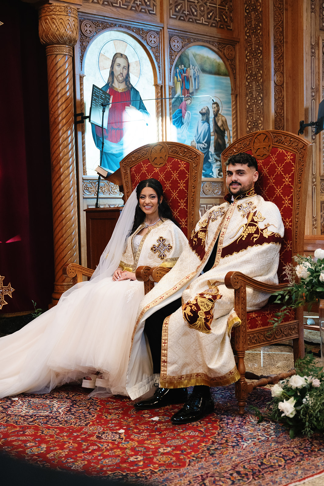 Bride and groom sit side by side on ornate red and gold thrones during their Coptic Orthodox wedding ceremony at St. Mary Church, wearing traditional embroidered ceremonial capes, surrounded by intricate wooden iconography and religious artwork.
