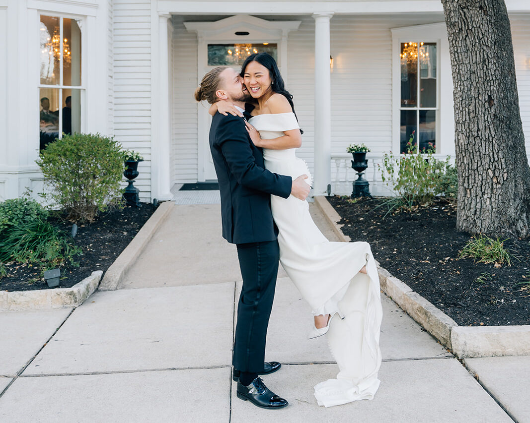 An elegant groom in a black tuxedo lifting his bride in a minimalist off-the-shoulder white wedding gown, sharing a joyful laugh in front of the historic Victorian facade of The Allan House in Austin, Texas.