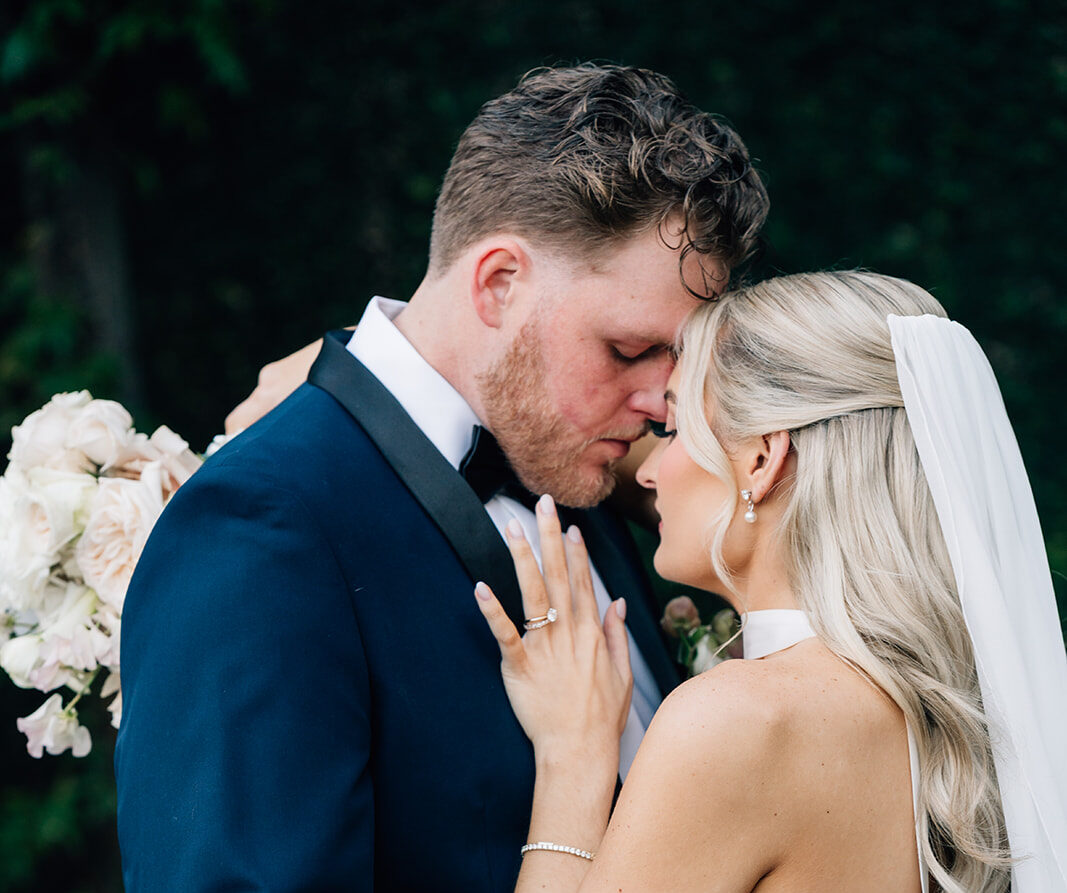 Close-up portrait of a bride and groom in an intimate embrace at The Bell Tower on 34th in Houston. The groom wears a navy tuxedo and the bride wears a sleek white wedding dress with a long veil.