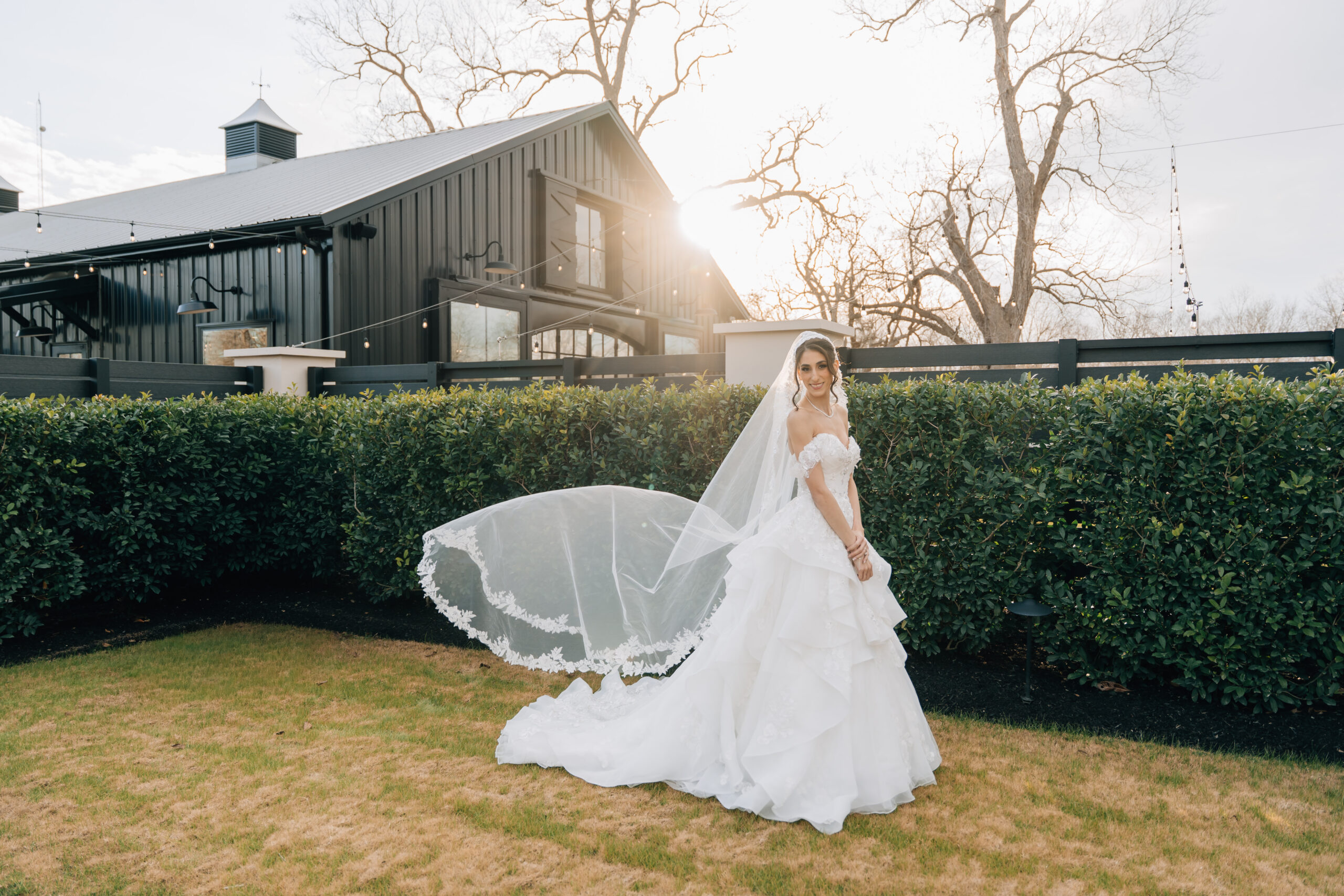 Editorial bridal portrait at Stables on the Brazos featuring a cathedral-length veil caught in the wind against a modern black barn.