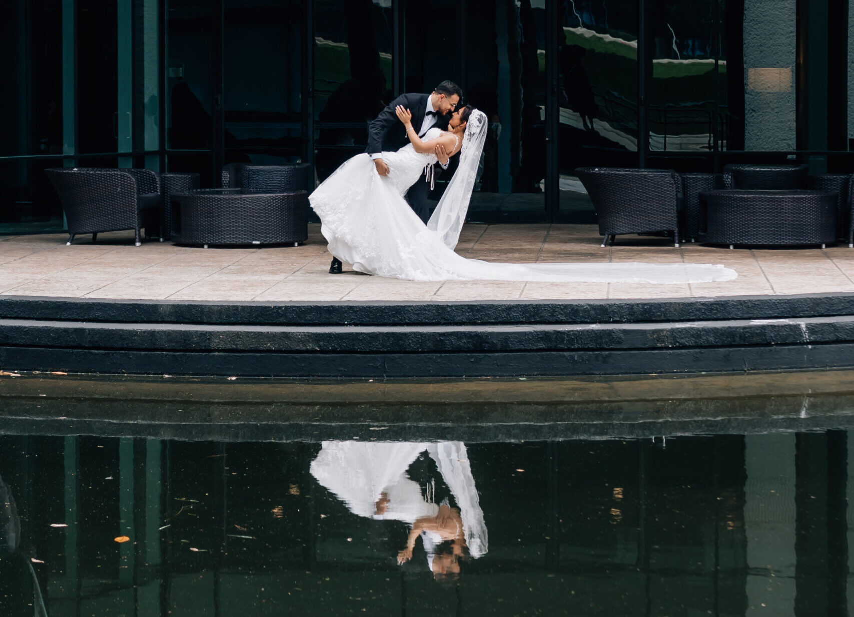Fine art wedding portrait of a bride and groom dipping by a modern glass building with a water reflection, captured by Houston wedding photographer Katia G Photography.